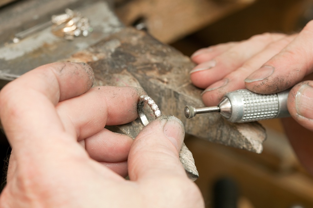 A jeweler, using a small flexible shaft grinder, is grinding the band of a diamond wedding ring. In the background are blurred rings on the workbench and the jeweler's hands are dirty